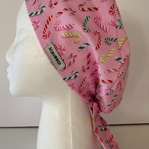 Puede incluir: Una gorra rosa con un estampado repetido de bastones de caramelo rojos, blancos y verdes. La gorra tiene una etiqueta que dice "SCRUBBED". La gorra se ata en la parte posterior con lazos largos.