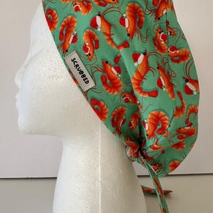 Puede incluir: Un gorro quirúrgico verde con un estampado repetido de gambas naranjas. La etiqueta blanca dice "SCRUBBED". El gorro se ata en la parte posterior y está diseñado para uso médico.