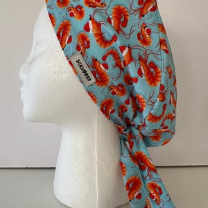 Puede incluir: Un gorro quirúrgico azul claro con un estampado repetido de camarones naranjas. El gorro tiene un lazo en la parte posterior y una pequeña etiqueta blanca que dice "SCRUBBED". Se muestra en una cabeza de maniquí blanca.