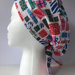 Puede incluir: Un gorro quirúrgico blanco con un estampado colorido de regalos envueltos en rojo, rosa, verde y azul. El gorro tiene un diseño con lazo en la parte trasera y una pequeña etiqueta que dice "Scrubbed".