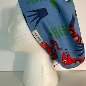 Peut inclure: Une toque de chirurgien bleue avec un motif répété de serpents rouges et verts, de feuilles et de formes abstraites. La toque porte une étiquette blanche avec l'inscription "SCRUBBED".