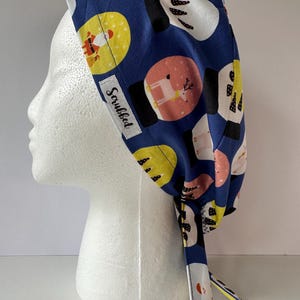 Puede incluir: Un gorro quirúrgico azul con un estampado festivo con Papá Noel, renos y formas abstractas en amarillo, blanco y rosa. El gorro tiene una etiqueta blanca con la palabra "Scrubbed" impresa.