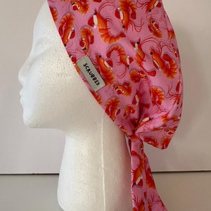 Puede incluir: Gorro quirúrgico rosa con un estampado de gambas naranjas y rojas. El gorro tiene un diseño con lazo en la parte trasera y una pequeña etiqueta que dice "SCRUBBED". Este accesorio médico está diseñado para profesionales de la salud.