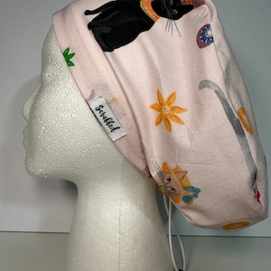 May include: A pink scrub hat with a white drawstring and a blue toggle. The hat features a repeating pattern of cats, flowers, and other whimsical designs. The text "Scrubbed" is printed on the hat.