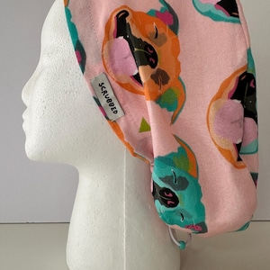 Könnte beinhalten: Eine rosa OP-Haube mit einem sich wiederholenden Muster aus Cartoon-Hundegesichtern in Orange, Türkis und Rosa. Die Haube hat einen Kordelzug und ein kleines Etikett mit der Aufschrift "SCRUBBED".