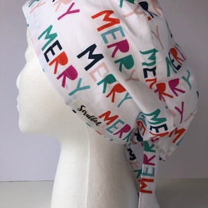 Puede incluir: Gorro quirúrgico blanco con la palabra "MERRY" en varios colores. El gorro está atado en la parte posterior y tiene la palabra "Scrubbed" en una pequeña etiqueta. El gorro está sobre una cabeza de maniquí blanca.