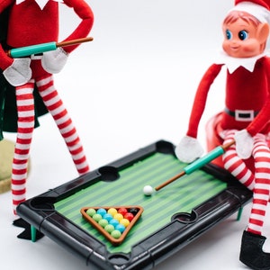 May include: Two elf figurines playing a miniature pool game. The elf on the left is holding a pool cue and the elf on the right is taking a shot. The pool table is green with a black frame and has a white ball and a set of colorful balls in a triangle.