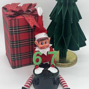 May include: A Christmas elf figurine with a red and white hat and suit, holding the number 67. A red plaid gift box with a glittery bow and a green paper Christmas tree are in the background. The elf has red and white striped legs and black shoes.