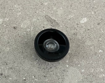 Könnte beinhalten: Eine schwarze Objektivkappe mit einer kleinen, klaren Linse in der Mitte.