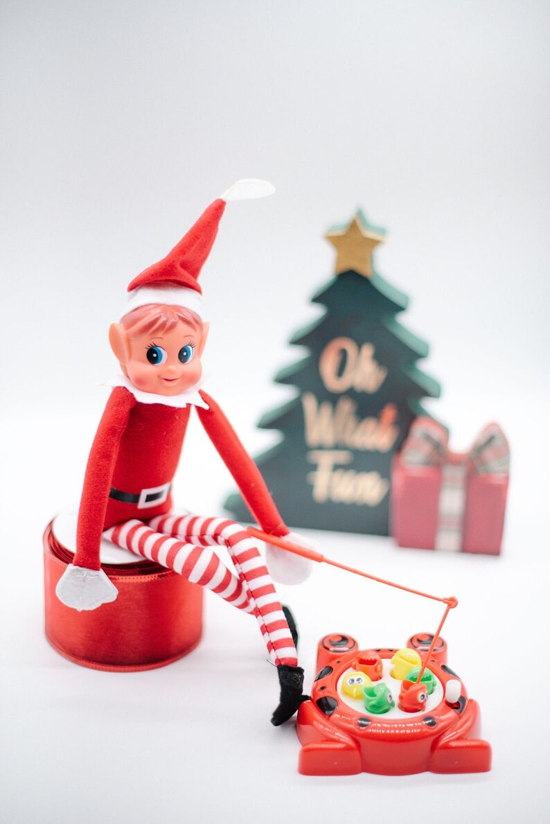 May include: A red elf doll with a white hat sits on a red spool and holds a fishing rod over a red plastic frog toy with colorful fish. The frog toy has a spinning wheel and the text "Oh What Fun" is visible on a green Christmas tree behind the elf.