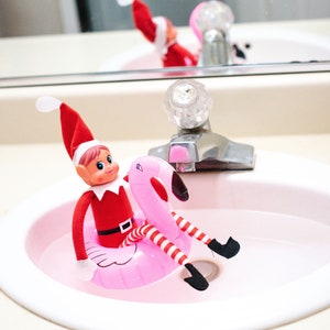 May include: A small elf doll wearing a red and white striped suit is sitting in a pink inflatable flamingo pool float in a white bathroom sink.