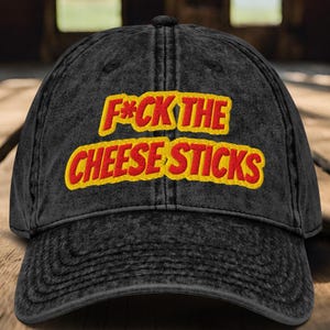 May include: A black, distressed baseball cap with the embroidered phrase "F*ck the Cheese Sticks" in yellow and red lettering. The cap has a curved brim and a classic six-panel design.