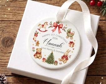 May include: A round white ceramic Christmas ornament featuring a red ribbon and illustrations of a rocking horse, teddy bear, rabbit, and tree. The text reads: Happy 1st Christmas 2026, Hannah, Love Nanny & Grandad. It includes a white satin ribbon.
