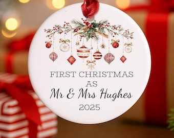 May include: A white, circular Christmas bauble with a red ribbon. The bauble displays a festive design with hanging ornaments and the text "FIRST CHRISTMAS AS Mr & Mrs Hughes 2025".