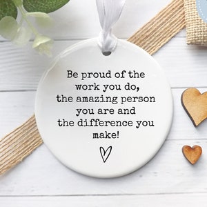 May include: White ceramic ornament with a black and white inspirational quote: "Be proud of the work you do, the amazing person you are and the difference you make!"  The ornament is decorated with a small heart and comes with two wooden heart embellishments.