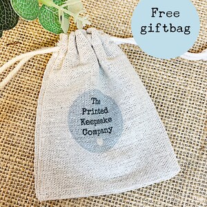 Pode incluir: Uma pequena bolsa de cord&atilde;o de cor branco-creme, feita de tecido natural. A bolsa tem um logotipo circular preto com o texto "The Printed Keepsake Company". A bolsa est&aacute; sobre uma superf&iacute;cie de juta marrom. Um c&iacute;rculo azul com o texto "Free giftbag" est&aacute; no canto superior direito da imagem.