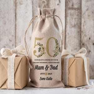 May include: A beige burlap wine bottle bag with a drawstring closure. The bag features a gold "50" with floral accents, and text that reads "CONGRATULATIONS ON YOUR GOLDEN WEDDING ANNIVERSARY Mum & Dad 1975 2025 Love Katie."