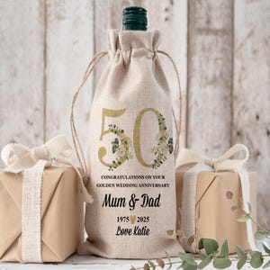 May include: A beige wine bottle bag with a drawstring closure. The bag features a gold "50" with floral accents, and text that reads "CONGRATULATIONS ON YOUR GOLDEN WEDDING ANNIVERSARY Mum & Dad 1975 2025 Love Katie."