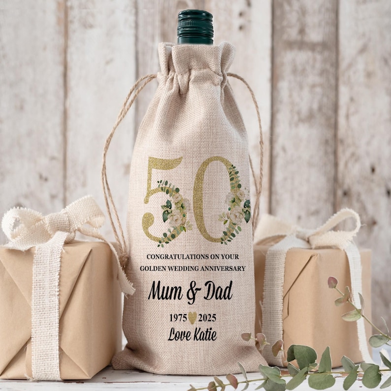 May include: A beige wine bottle bag with a drawstring closure. The bag features a gold "50" with floral accents, and text that reads "CONGRATULATIONS ON YOUR GOLDEN WEDDING ANNIVERSARY Mum & Dad 1975 2025 Love Katie."