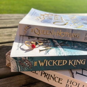 May include: Two paperback books stacked on top of each other. The top book is titled "The Queen of Nothing" and the bottom book is titled "The Wicked King". Both books have a blue and green cover with gold lettering. A gold ring with a red and black stone is on top of the books.