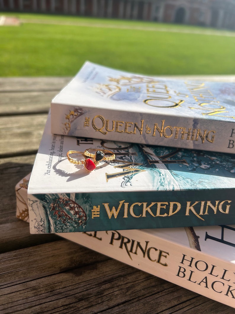 May include: Two paperback books stacked on top of each other. The top book is titled "The Queen of Nothing" and the bottom book is titled "The Wicked King". Both books have a blue and green cover with gold lettering. A gold ring with a red and black stone is on top of the books.