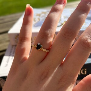 May include: A gold ring with a black heart-shaped stone. The ring is on a finger.