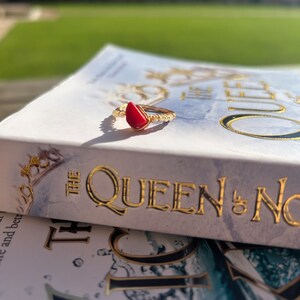 May include: A gold ring with a red gemstone in the shape of a teardrop. The ring is resting on a book titled "The Queen of Nothing".