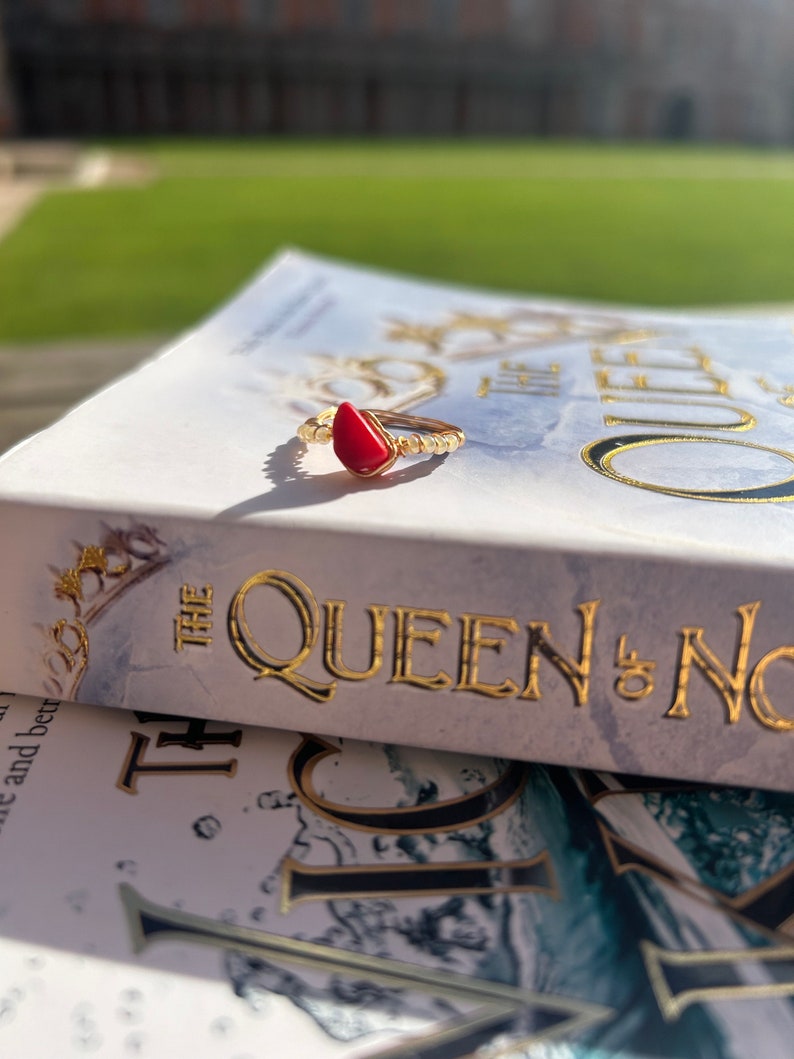 May include: A gold ring with a red gemstone in the shape of a teardrop. The ring is resting on a book titled "The Queen of Nothing".