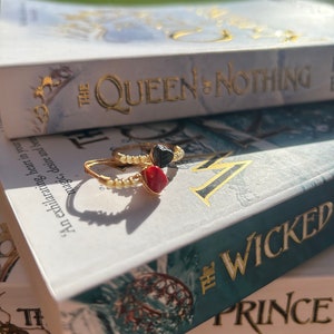 May include: Two gold rings with black and red stones on top of a book titled "The Wicked Prince" with the words "The Queen of Nothing" on the book above it.