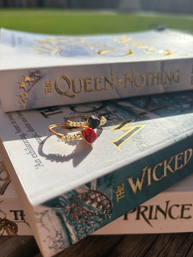 May include: Two gold rings with black and red stones on top of a book titled "The Wicked Prince" with the words "The Queen of Nothing" on the book above it.