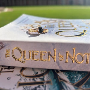 May include: A gold ring with a black heart-shaped stone. The ring is sitting on a book titled "The Queen & Not".