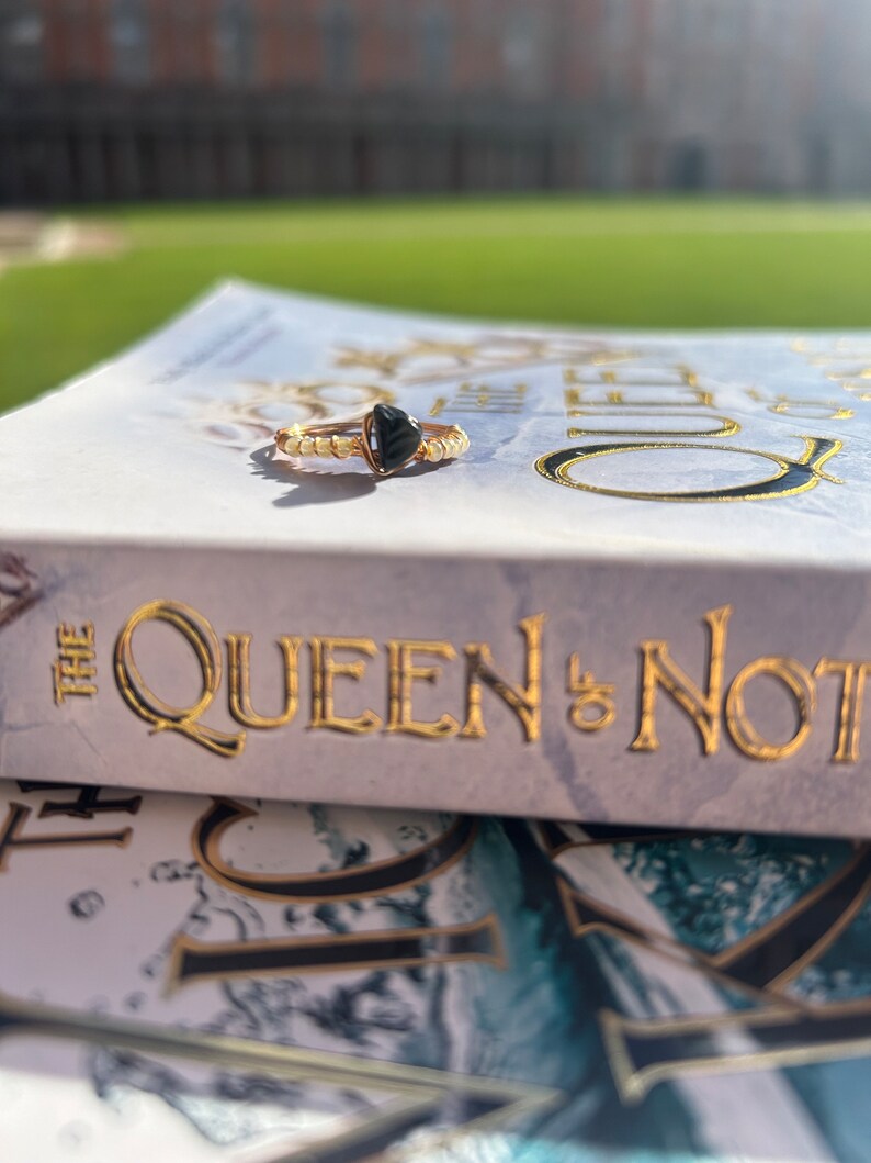 May include: A gold ring with a black heart-shaped stone. The ring is sitting on a book titled "The Queen & Not".