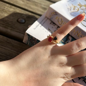 May include: A gold ring with two heart-shaped stones, one red and one black, on a finger. The ring is on a hand resting on a book with the title "The Queen of the North" visible.