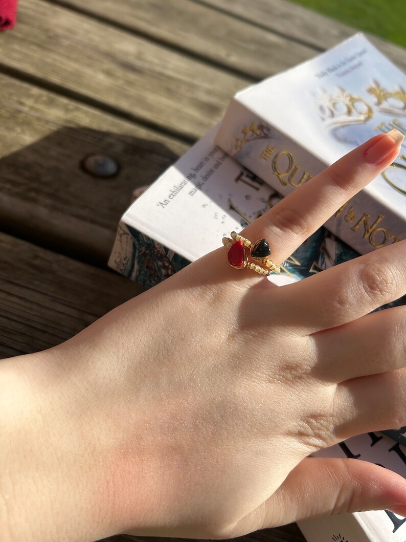 May include: A gold ring with two heart-shaped stones, one red and one black, on a finger. The ring is on a hand resting on a book with the title "The Queen of the North" visible.