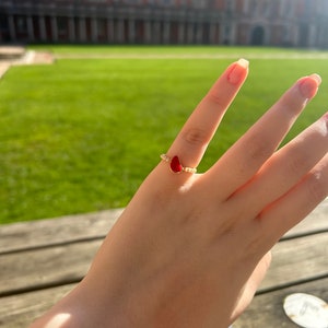 May include: A gold ring with a red teardrop-shaped stone and small white beads around the band.