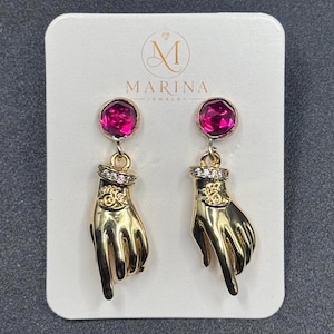 May include: A pair of gold-toned earrings on a white display card labelled MARINA JEWELRY. Each earring features a round, faceted fuchsia crystal stud connected to a gold-toned hand charm with a small eye symbol and a band of tiny clear stones.