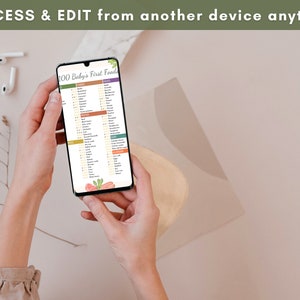 May include: A person is holding a smartphone displaying a digital checklist titled "100 Baby's First Foods". The checklist is organized into categories, including fruits, vegetables, grains, and proteins. The phone is on a light brown surface.