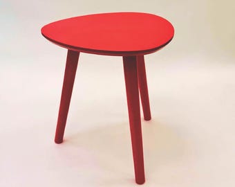May include: A small, red, three-legged side table. The tabletop is a rounded triangle shape. The legs are cylindrical and angled outward. The table is painted a solid, bright red color. The table is suitable for a living room or bedroom.