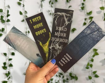 May include: Four rectangular bookmarks featuring dark themes. One displays 'I NEED MORE BOOKS' with a green claw, another 'READ MORE HORROR' with skeletal imagery, one says 'HORROR STORIES ARE MY HAPPY PLACE', and the last reads 'THERE IS A STRANGE COMFORT IN BEING ALONE'.
