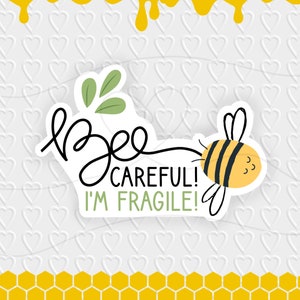 Può includere: Un adesivo bianco con un'illustrazione di un'ape e il testo "Bee careful! I'm fragile!" su uno sfondo bianco con un motivo di cuori bianchi e un motivo a nido d'ape giallo nella parte inferiore.