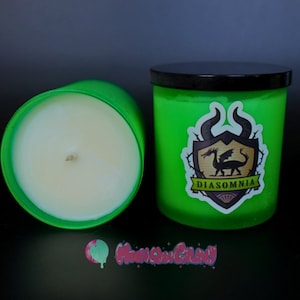 May include: Two green scented candles with black lids. One candle is open, revealing a white wax interior. The other candle has a sticker with a dragon and the word "DIASOMNIA". The candles are on a black surface.