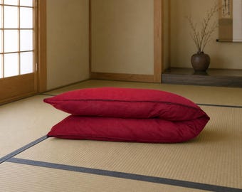 May include: A rectangular floor cushion in a deep red fabric with a dark, thin piping detail along the edges. The cushion is folded in half, creating a stacked appearance with a soft, textured surface visible on the exterior of the fabric.