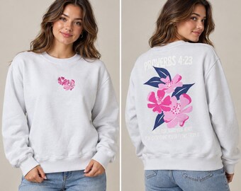 May include: A light gray crewneck sweatshirt featuring a small pink floral graphic on the front chest. The back displays a large floral design in shades of pink and dark blue with the text Proverbs 4:23 and Above all else guard your heart.