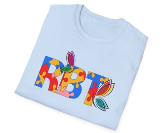 May include: Light blue t-shirt with the word "RBT" spelled out in puzzle pieces. The puzzle pieces are in various colors, including red, yellow, blue, and green. There are two butterfly-shaped designs on the shirt, one above the word "RBT" and one to the right of the word.