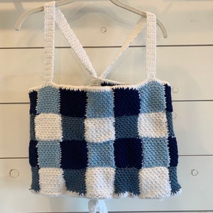 Pode incluir: Um top de crochê branco com um padrão xadrez azul e branco. O top tem alças finas que cruzam nas costas.