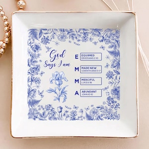 May include: A square, white ceramic trinket dish with a gold rim. The dish features a blue floral design and the text "God Says I am" with the words "Equipped, Made New, Merciful, Abundant" and corresponding Bible verses.