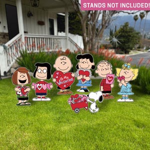 Op de afbeelding: Tuinversieringen met stripfiguren die Valentijnsdag-harten vasthouden. De figuren zijn onder meer Charlie Brown, Lucy, Linus, Sally en Snoopy met een rode wagen. De tekst "Be My Valentine" is zichtbaar op een van de harten.