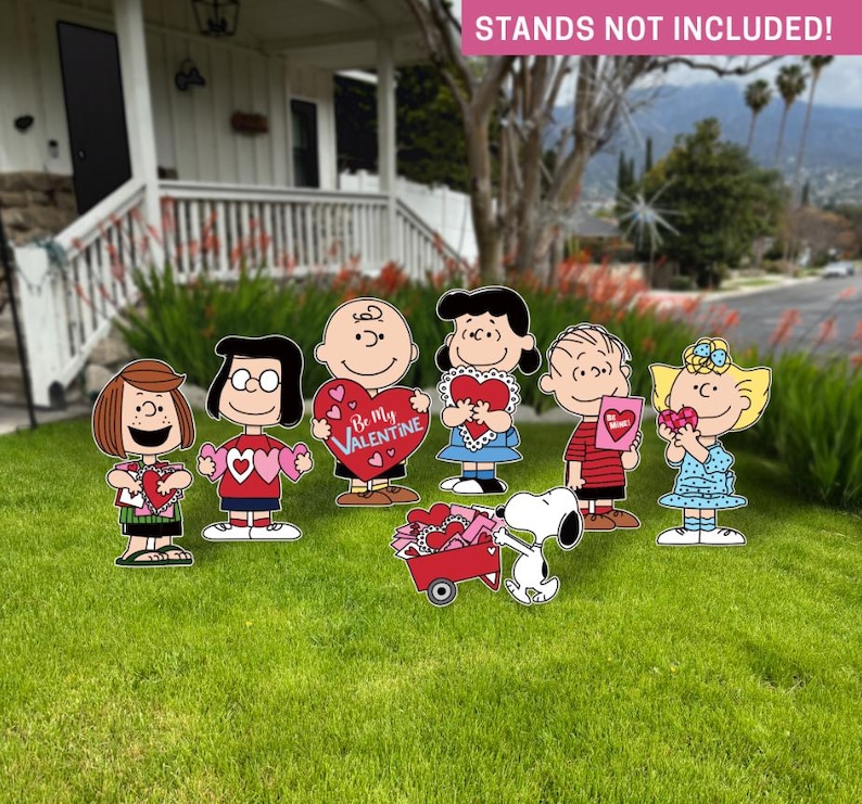 Op de afbeelding: Tuinversieringen met stripfiguren die Valentijnsdag-harten vasthouden. De figuren zijn onder meer Charlie Brown, Lucy, Linus, Sally en Snoopy met een rode wagen. De tekst "Be My Valentine" is zichtbaar op een van de harten.