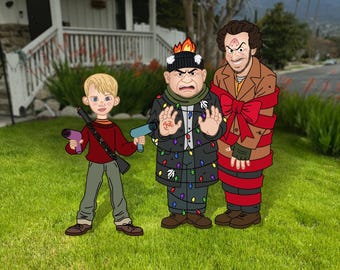 May include: Cartoon cutouts of three characters from a holiday movie, standing on green grass in front of a house. One character holds a toy gun and a ray gun. Another is wrapped in red ribbon. The third is wearing a coat decorated with Christmas lights. Text at the top reads "STANDS NOT INCLUDED!"
