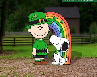 Puede incluir: Snoopy y Charlie Brown vestidos de verde y con un sombrero de duende se paran frente a un arcoíris. "Stands not included" está escrito en la parte superior de la imagen.